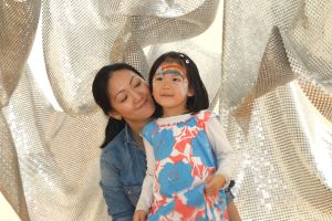 A mother and daughter stand smiling in front of a metallic backdrop, with the young girl sporting a rainbow painted on her forehead, suggesting they’re enjoying a fun face-painting event.