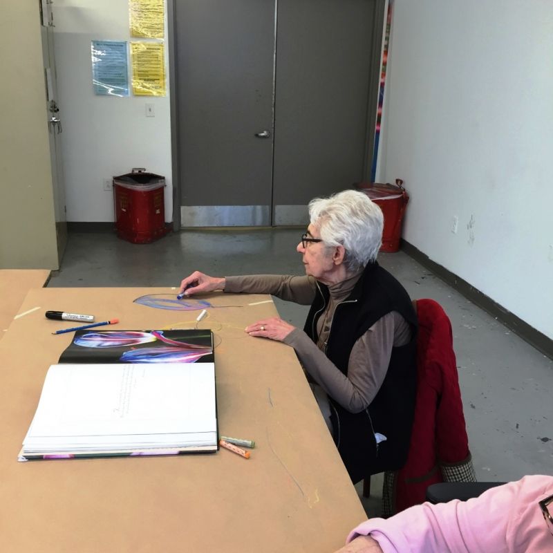 A woman at a desk with an open book and art supplies in front of her.