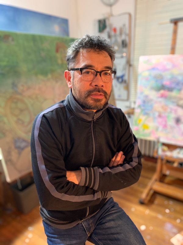 Hiro Sakaguchi sits in studio with crossed arms and artwork in background