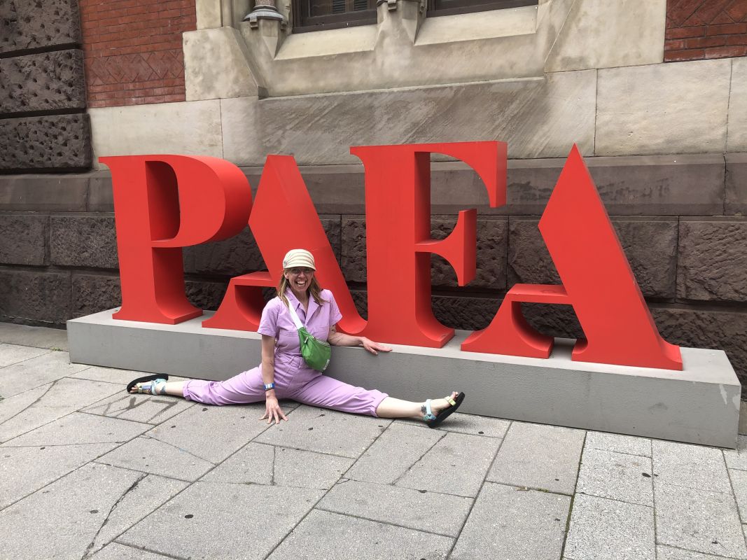 Kristy Jane smiles, doing the splits in front of a large PAFA sign