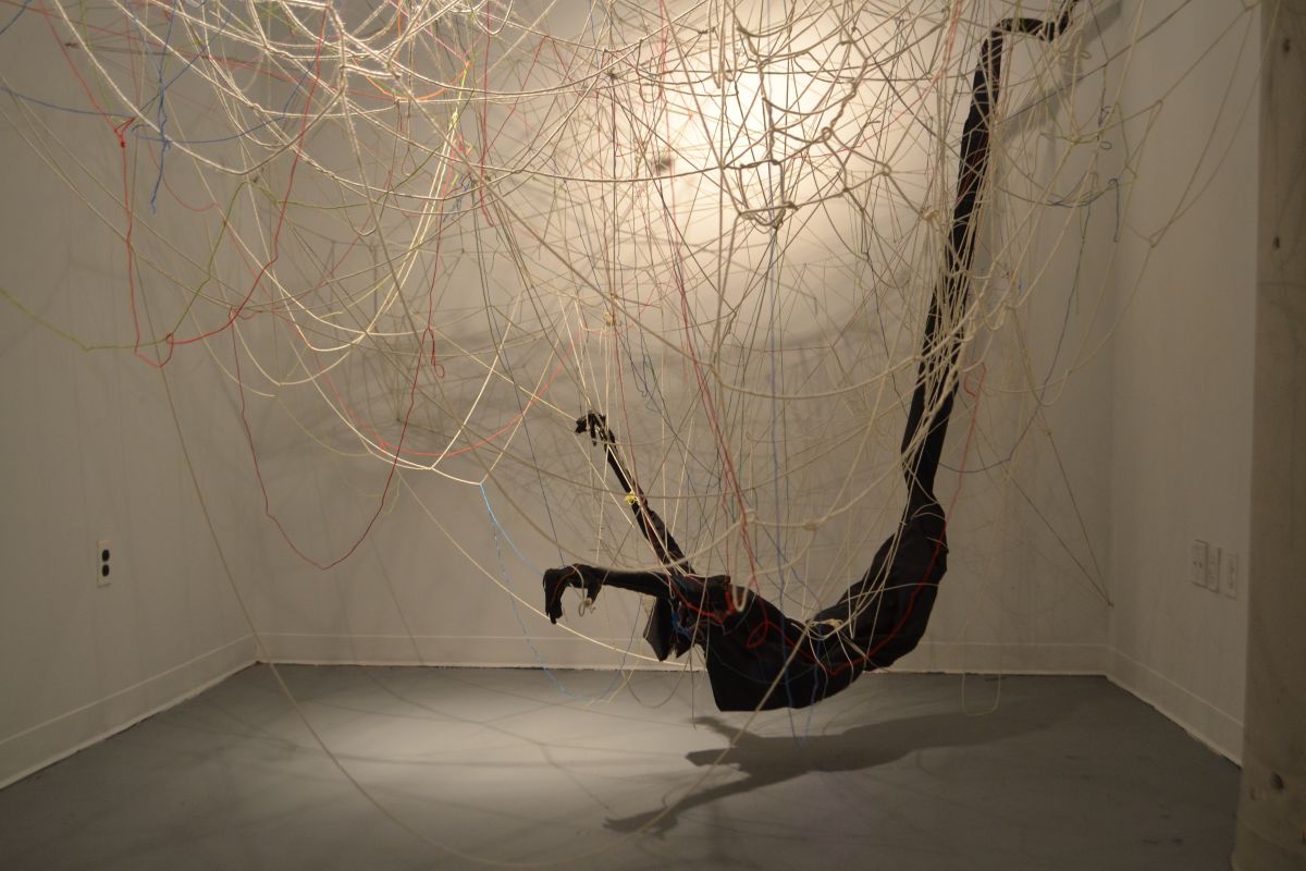 Humanoid figure is suspended in room by tangle of threads
