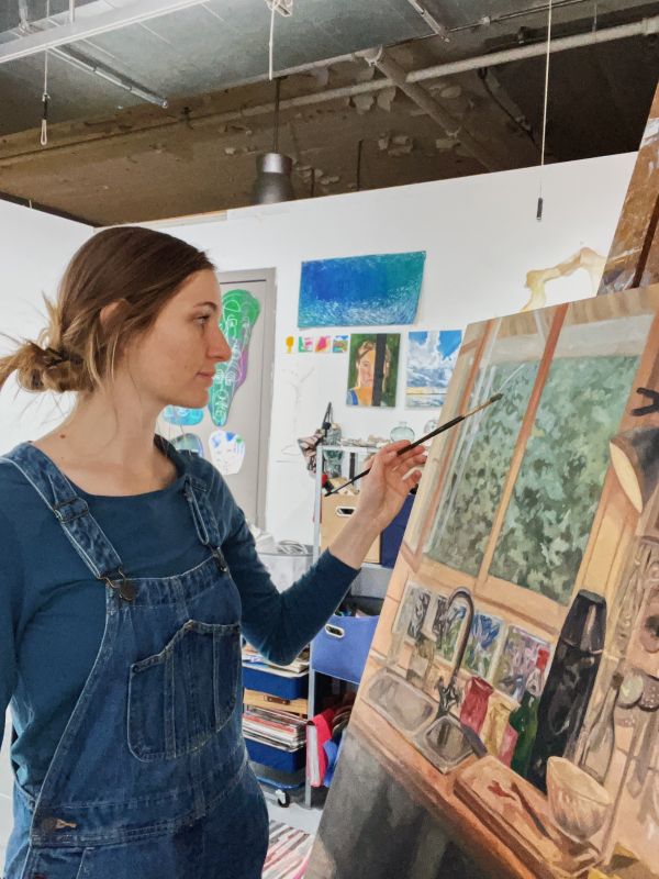 Woman in blue outfit holds paintbrush to easel in studio