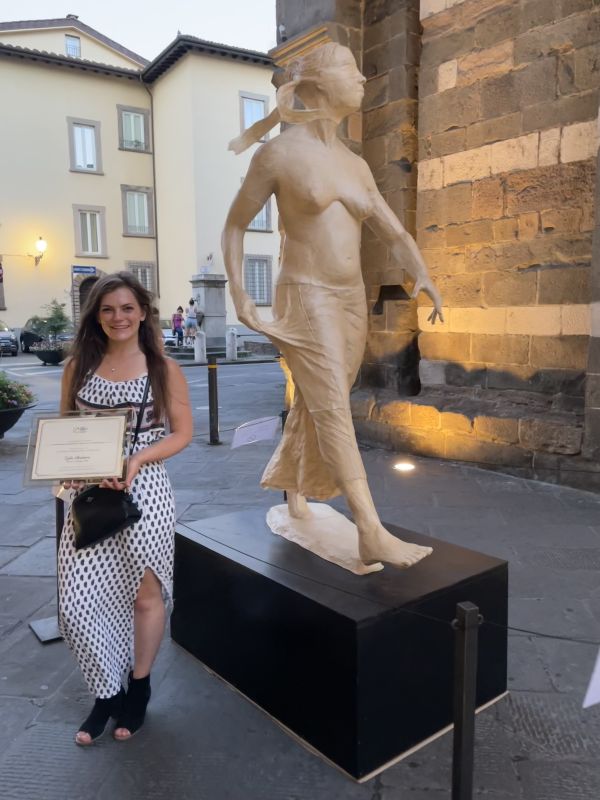 A woman wearing a black and white spotted dress holds a certificate, standing in front of figure sculpture