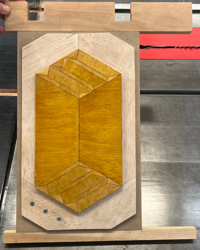 a piece of wood inlay or marquetry being worked on, likely using a jig on a saw table. 