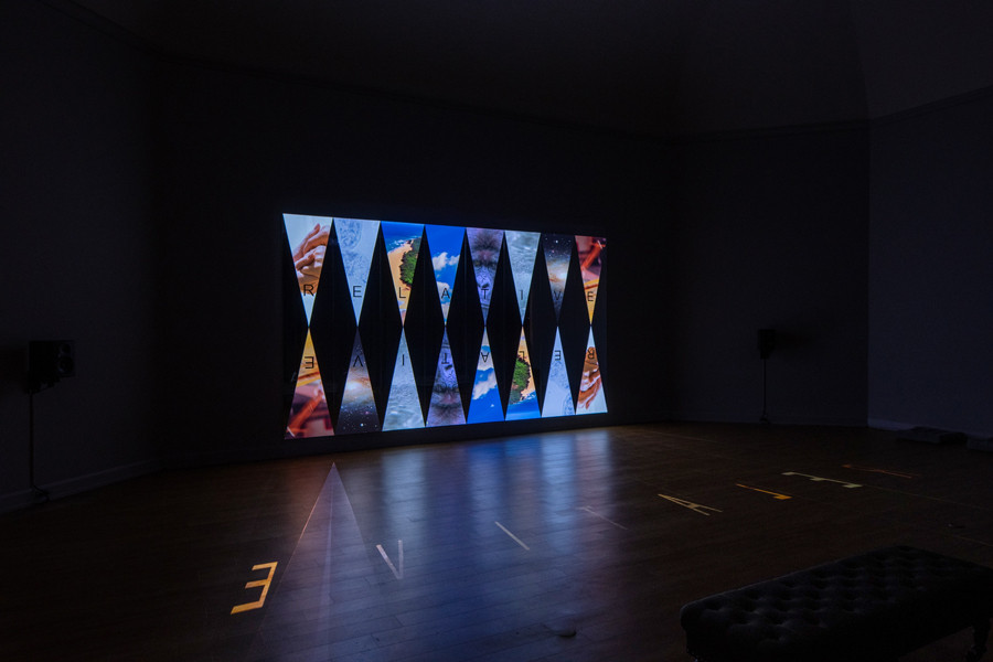 A dark room with a vibrant digital art display featuring triangular panels on the wall. The panels show abstract patterns and landscapes, reflecting on the wooden floor.