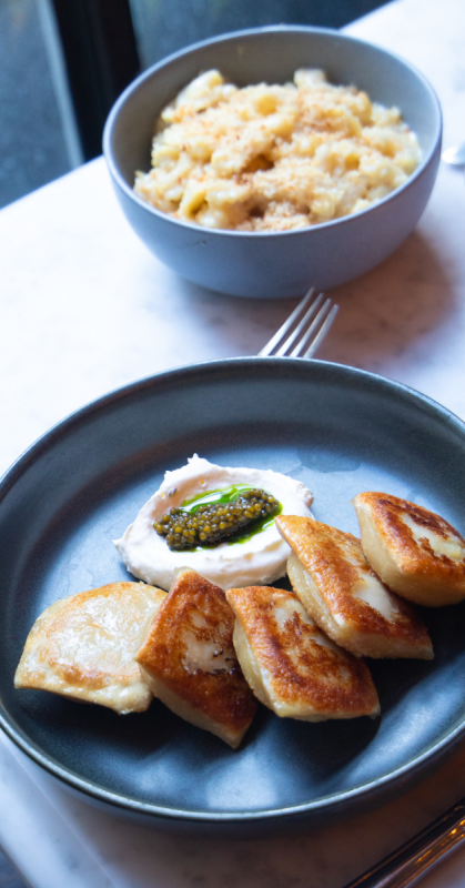 one sentence descriptive alt textA dark plate holds five pan-seared, golden-brown pierogi arranged alongside a dollop of white cream topped with green caviar, while a grey bowl filled with macaroni and cheese sits blurred in the background.