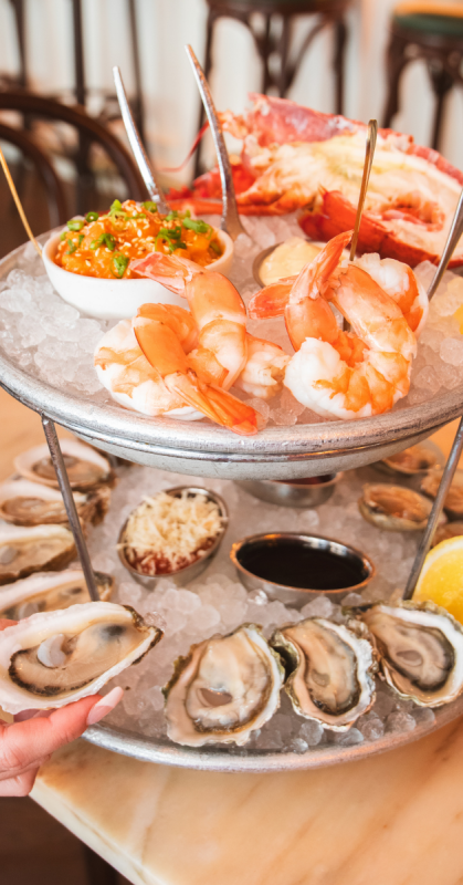 A two-tiered metal seafood tower filled with ice showcases fresh oysters on the bottom level and large poached shrimp, ceviche, and cooked lobster on the top level.