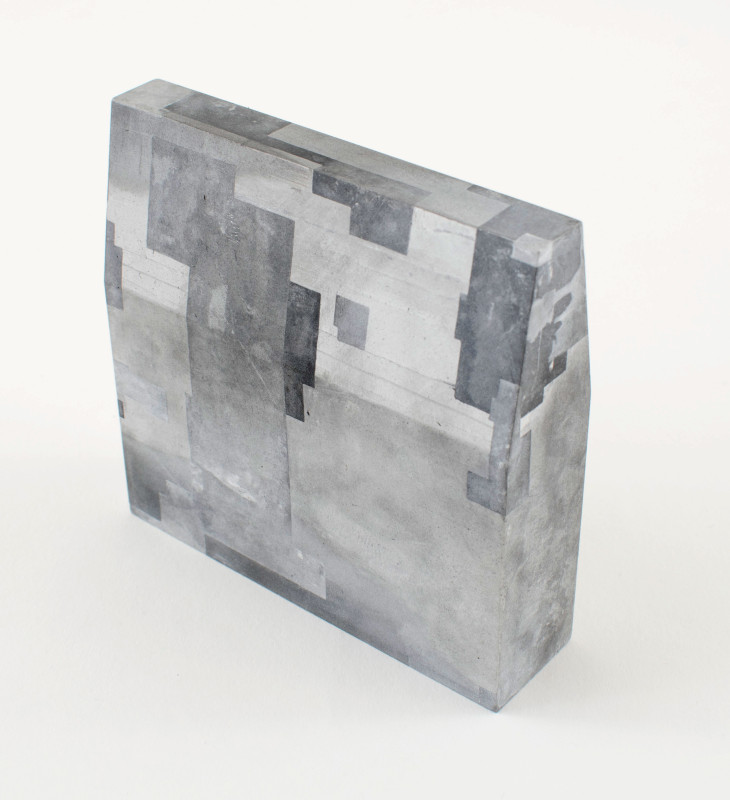 A wedge-shaped concrete sculpture with a heavily textured, monochromatic gray surface featuring layered, geometric block patterns reminiscent of brutalist architecture.