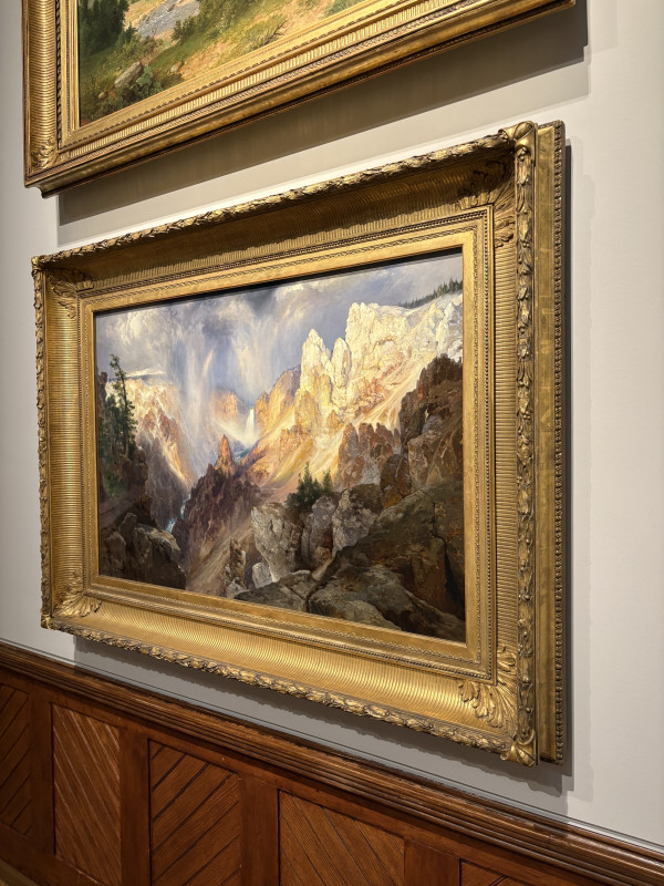 An oil painting with an ornate gold frame captures a dramatic canyon landscape under a stormy sky, where sunbeams pierce through dark clouds to illuminate yellow and white rocky cliffs.