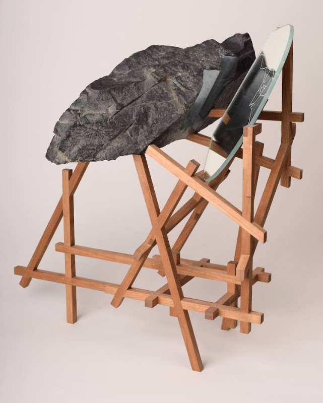 A modern sculptural artwork features a large, dark faux-stone rock and an oval mirror precariously balanced atop an intricate, geometric framework of crisscrossing light wooden dowels.