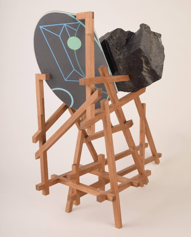 A complex, multi-tiered wooden scaffolding structure supports a large circular panel decorated with geometric designs and a large, dark, irregular rock resting against it.