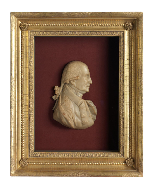A profile wax relief portrait of George Washington, depicted with his hair tied in a queue, is mounted on a red velvet background inside an ornate, rectangular gold shadowbox frame.