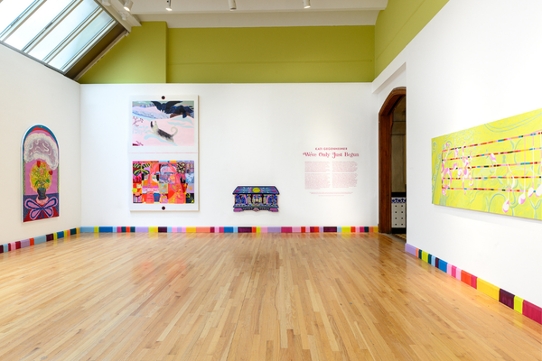 An art gallery exhibition room features colorful, vibrant paintings displayed on white walls above a light wood floor and a multi-colored baseboard block pattern.