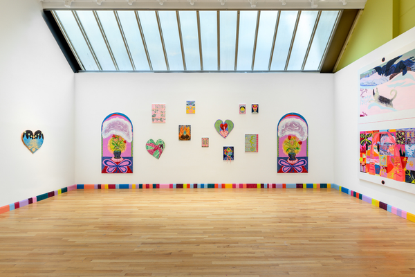 An art gallery installation view features brightly colored paintings and heart-shaped mixed-media works arranged on white walls under a large skylight, accented by a multicolored, block-patterned baseboard bordering the hardwood floor.