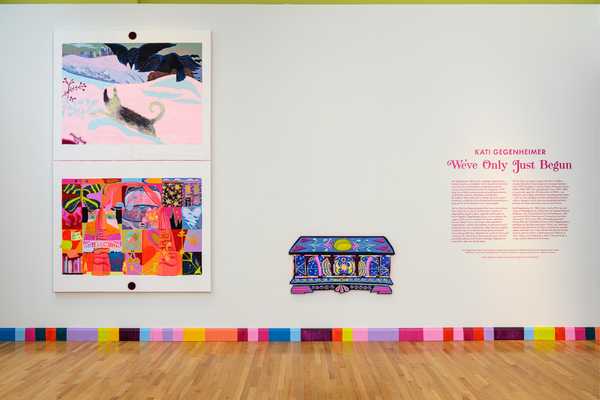 An art exhibition gallery wall displays two colorful paintings stacked vertically on the left, a vibrant chest-shaped artwork in the center, and descriptive pink wall text titled "We've Only Just Begun" on the right.