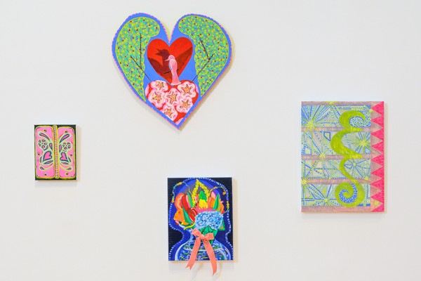 Four colorful, patterned paintings in various shapes, including a large heart, are arranged on a white gallery wall.
