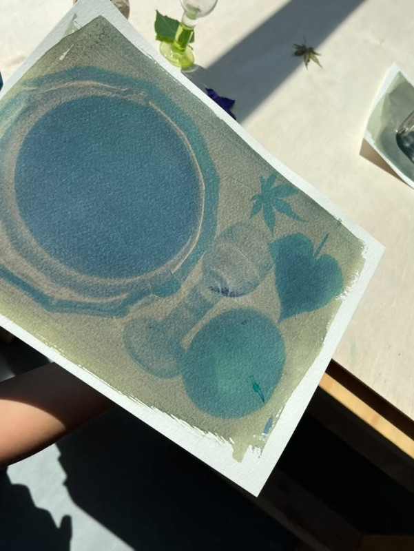 A hand holds up an exposed cyanotype print showing the blue-green silhouettes of a plate, a glass, a piece of fruit, and leaves before it has been washed.