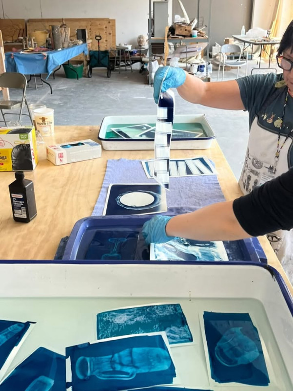 People working at a wooden table in an art studio, rinsing and developing blue-and-white cyanotype photographic prints in shallow water trays.