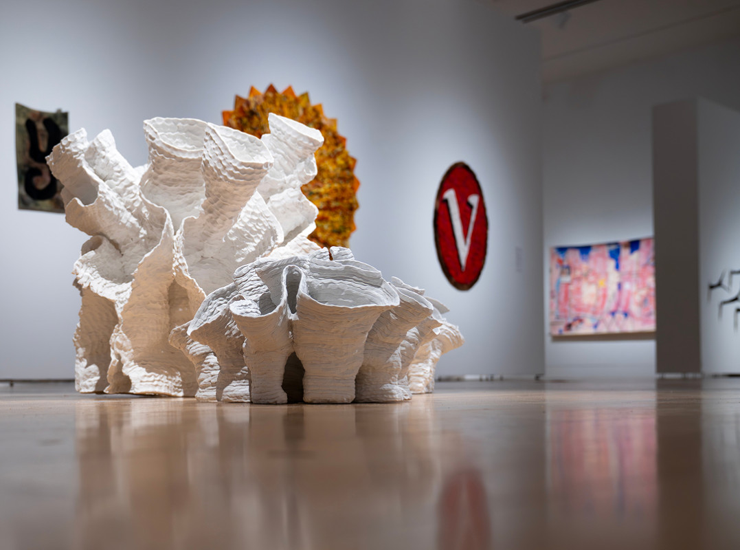 An organic, multi-tiered white ceramic sculpture resembling a cluster of textured coral sits on a polished gallery floor, surrounded by abstract paintings and artworks in a brightly lit museum exhibition space.
