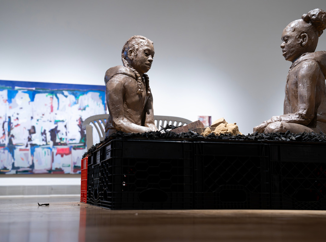 A low-angle shot of two textured clay sculptures of Black youths facing each other on a base of stacked black crates inside an art gallery with a large abstract painting in the background.