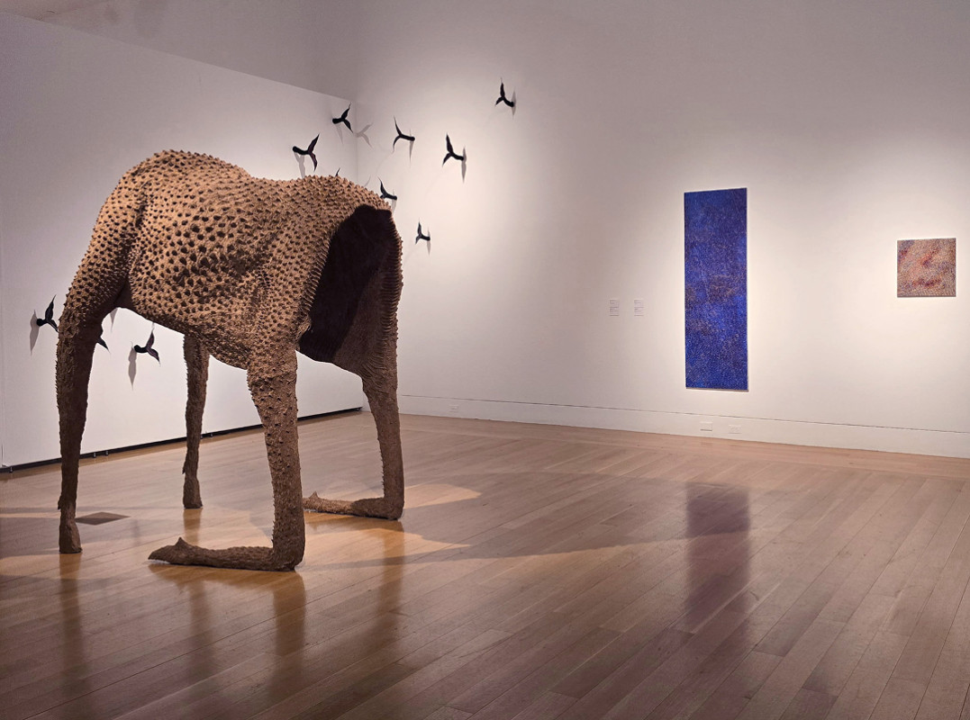 A large, hollowed-out, headless animal sculpture with a dimpled, textured surface stands on four legs in an art gallery, positioned near a flock of small sculpted birds mounted on the background wall alongside a vertical blue monochrome painting.