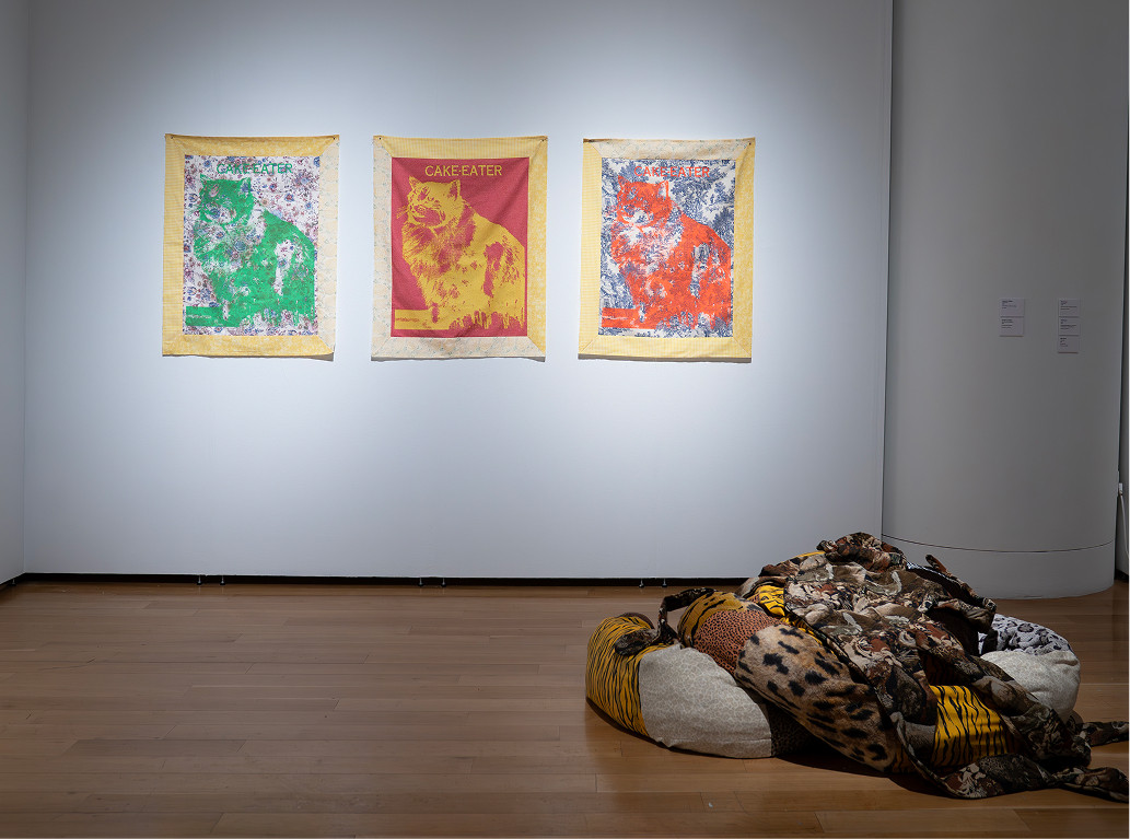 An installation view of a contemporary art exhibition featuring three vibrant, wall-hung feline portraits labeled "CAKE EATER" above a plush, patterned fabric floor sculpture.