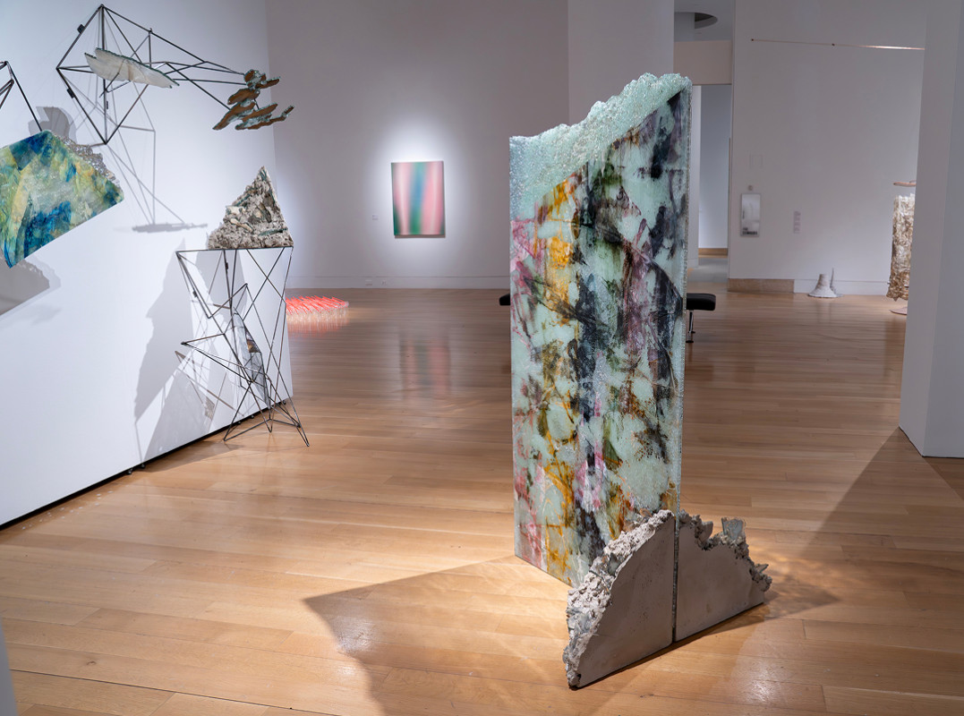 An upright, rectangular mixed-media sculpture with a swirling marbleized pattern stands on a concrete base inside a brightly lit art gallery, surrounded by wireframe wall structures and polished hardwood floors.