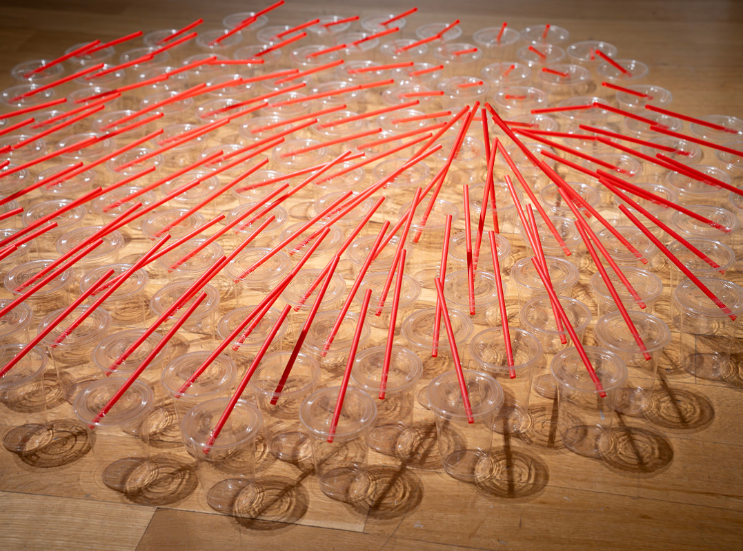 The image shows a contemporary sculpture installation composed of numerous clear plastic cups arranged in a circular floor grid, pierced and connected by radiating bright red plastic drinking straws.