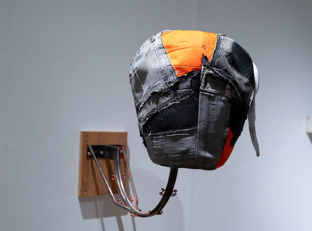 A mixed-media sculpture featuring a large, head-like form constructed from stitched patches of grey, black, and orange fabric, which is mounted to a wall using curved metal rods attached to a wooden base.