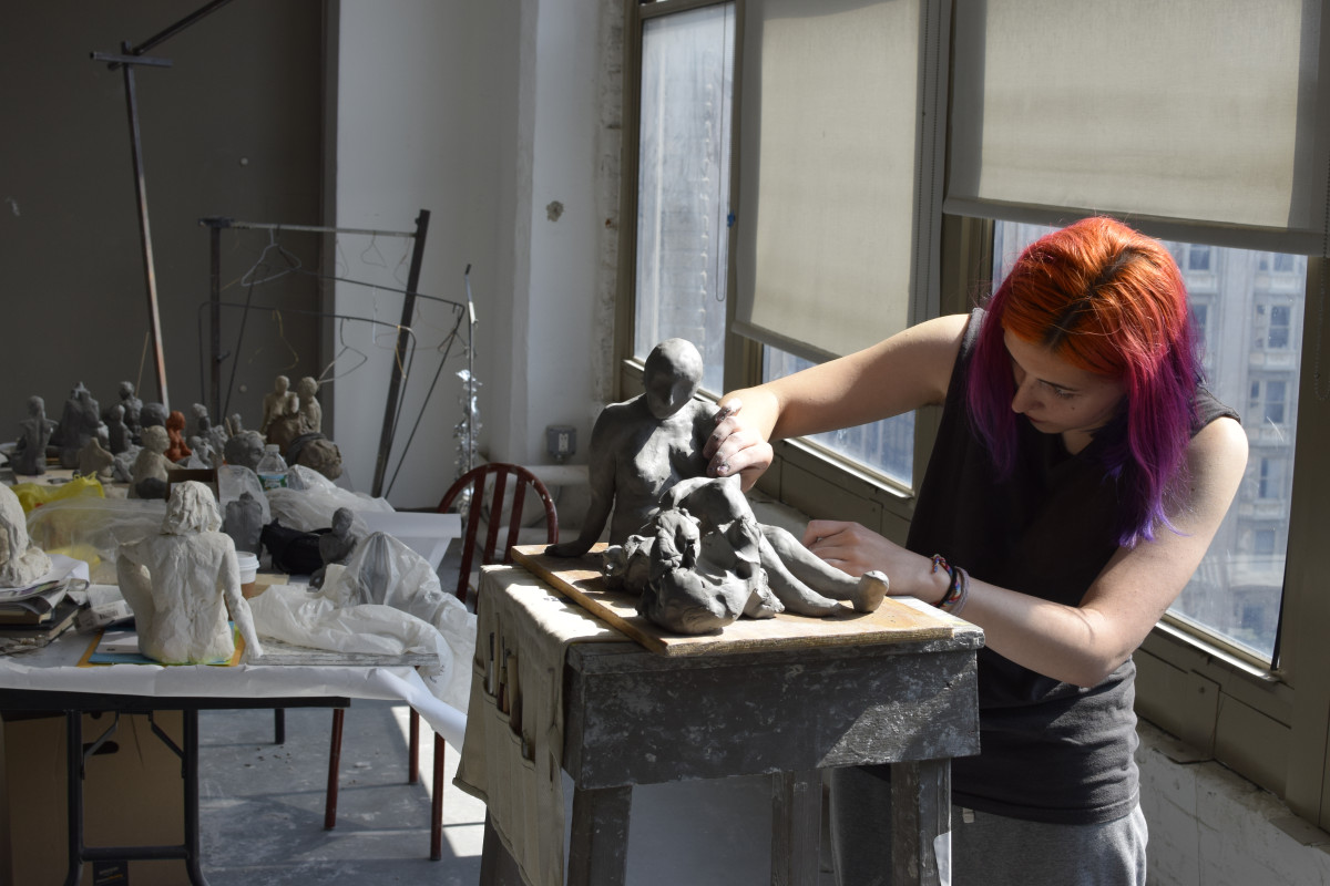 An artist with vibrant orange and purple hair carefully refines a clay sculpture of two figures on a wooden stand inside a sunlit studio filled with artwork.