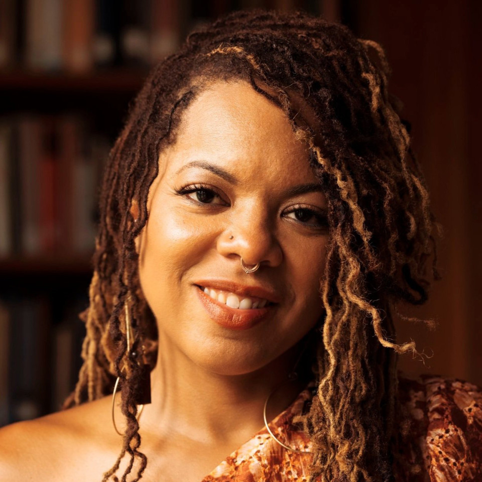 Bio photo of Airea D. Matthews a brown-skinned person, with long wavy brown hair, smiling and tilting their head to the right (a bit) 