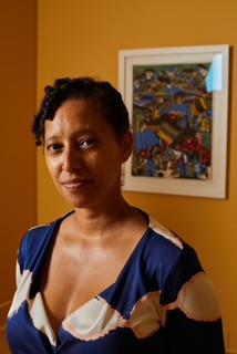 Bio photo of Kimberli Gant, brown woman with hair pulled back and wearing a blue with white flower print summer dress. 
