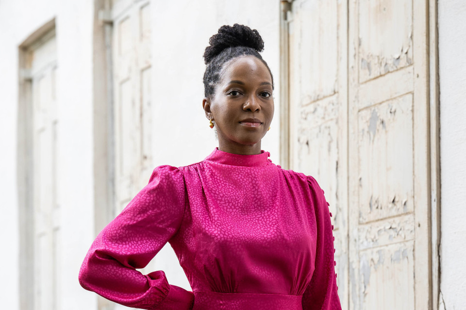 bio photo if Imani Perry, a person with brown skin, braided hair gathered in bun on top of their head, and wearing a bright pink blouse. 