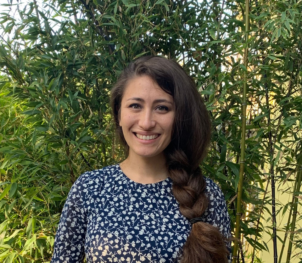 Bio photo of Grace Yasumura, smiling, light-skinned with long brown hair gathered in a thick braid draped over their shoulder, wearing a printed blue and white top. 