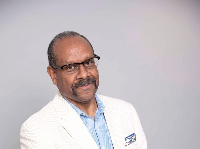 Bio photo of Allan L. Edmunds a brown-skinned man with a mustache. He is wearing glasses and a white suite jacket with a light blue button-down shirt. 