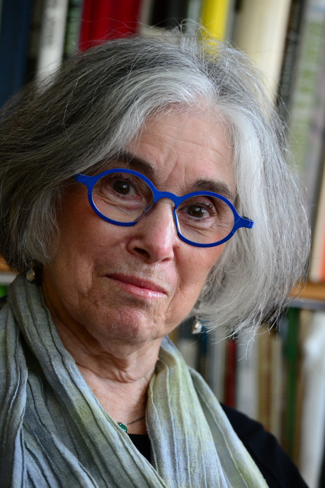 Bio photo of Ruth Fine, light skinned woman with salt and pepper hair, wearing round dark blue classes and wearing a blue-gray scarf. 