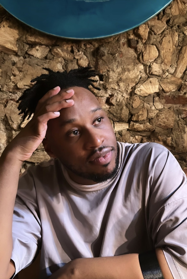 Bio photo of Marcellus who has brown skin with small dreadlocks and short beard posing with left arm resting in front of the body and right hand holding forehead. Marcellus is looking left and up slightly, with a relaxed face. 