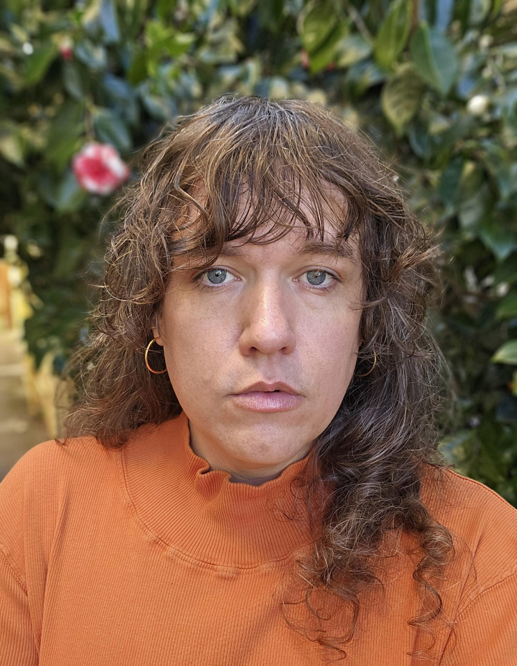 A person with wavy brown hair and bangs looks directly at the camera while wearing a burnt-orange, mock-neck ribbed shirt against a backdrop of dark green foliage.