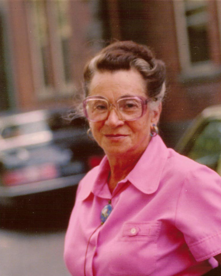 An older woman with grey-streaked brown hair and large pink-rimmed glasses wears a bright pink button-down shirt outdoors.