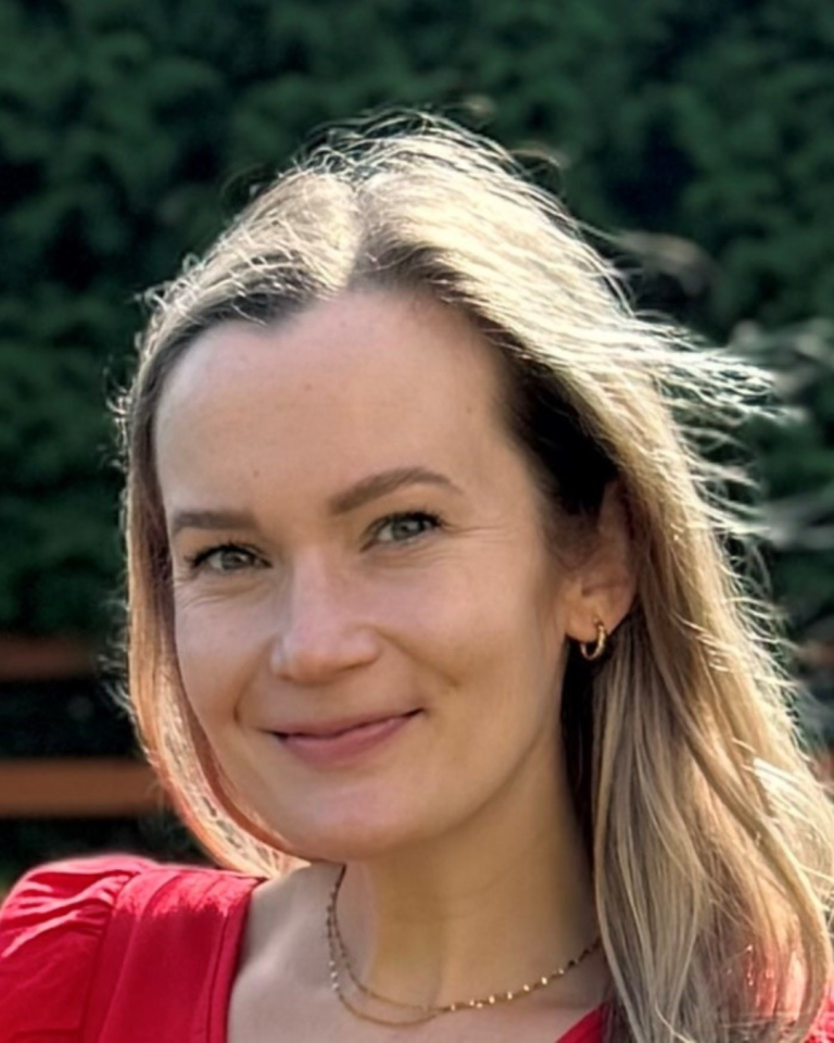 A headshot of a smiling Julie Nolan with blonde hair, wearing a red top, a gold necklace, and small gold hoop earrings against a soft, green background.