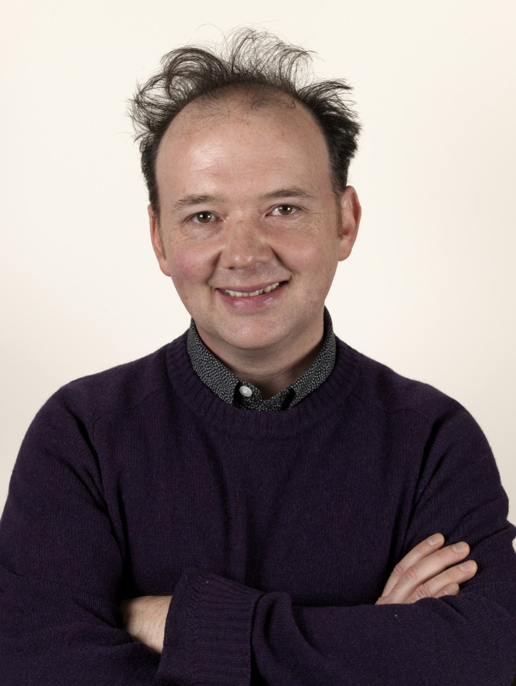 A man with thinning dark hair and a light complexion smiles for a portrait, wearing a purple crewneck sweater over a patterned collared shirt while standing with his arms crossed.
