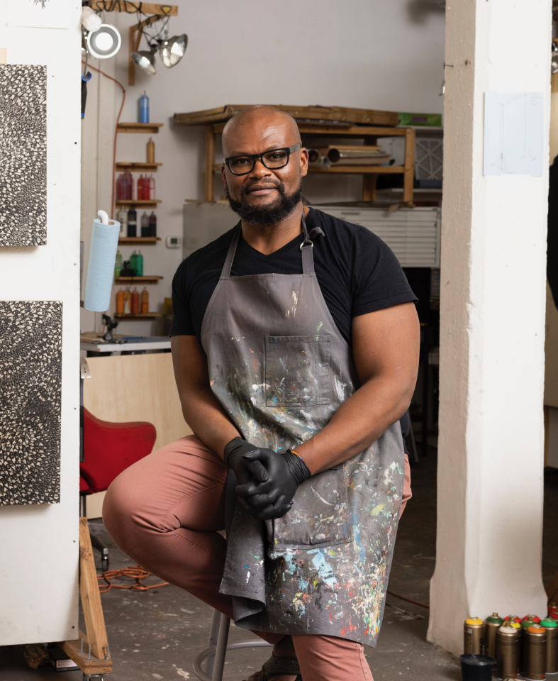 A bald Black man with a beard and glasses sits in an art studio, wearing a paint-splattered grey apron over a black t-shirt and pink trousers.