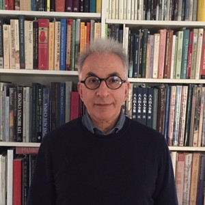An older man with gray hair and round glasses stands in front of floor-to-ceiling bookshelves filled with diverse titles.