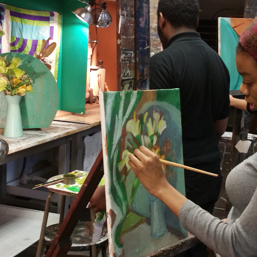 An artist's hand carefully applies a brushstroke to an impressionistic floral painting on a canvas in a brightly lit studio filled with other painters and still-life arrangements.