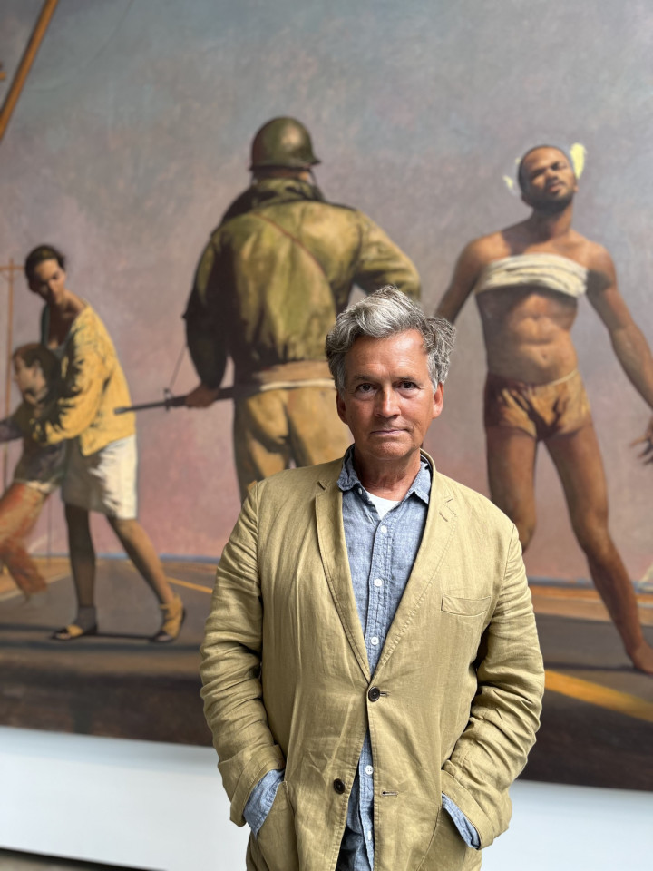 A man in a beige jacket stands in front of a large painting depicting historical scenes with soldiers and captives, evoking a somber and reflective tone.
