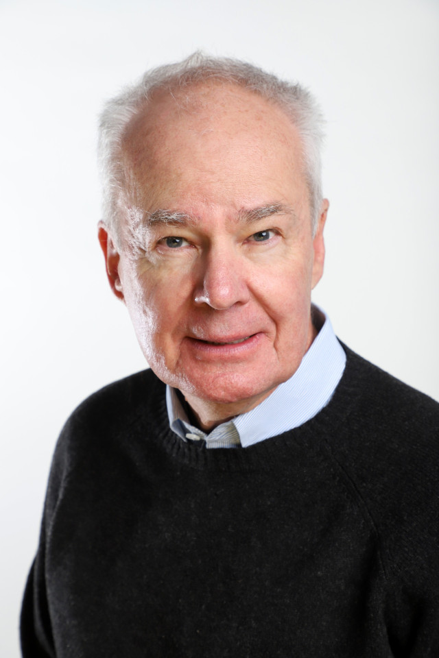 Elderly man with short white hair wearing a dark sweater over a light blue shirt, smiling slightly against a plain white background.