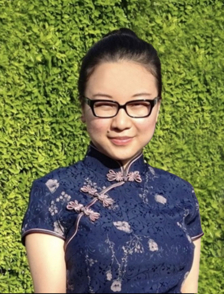 A woman stands against a lush green hedge, wearing glasses and a navy blue cheongsam with floral patterns. She smiles softly, conveying a calm mood.