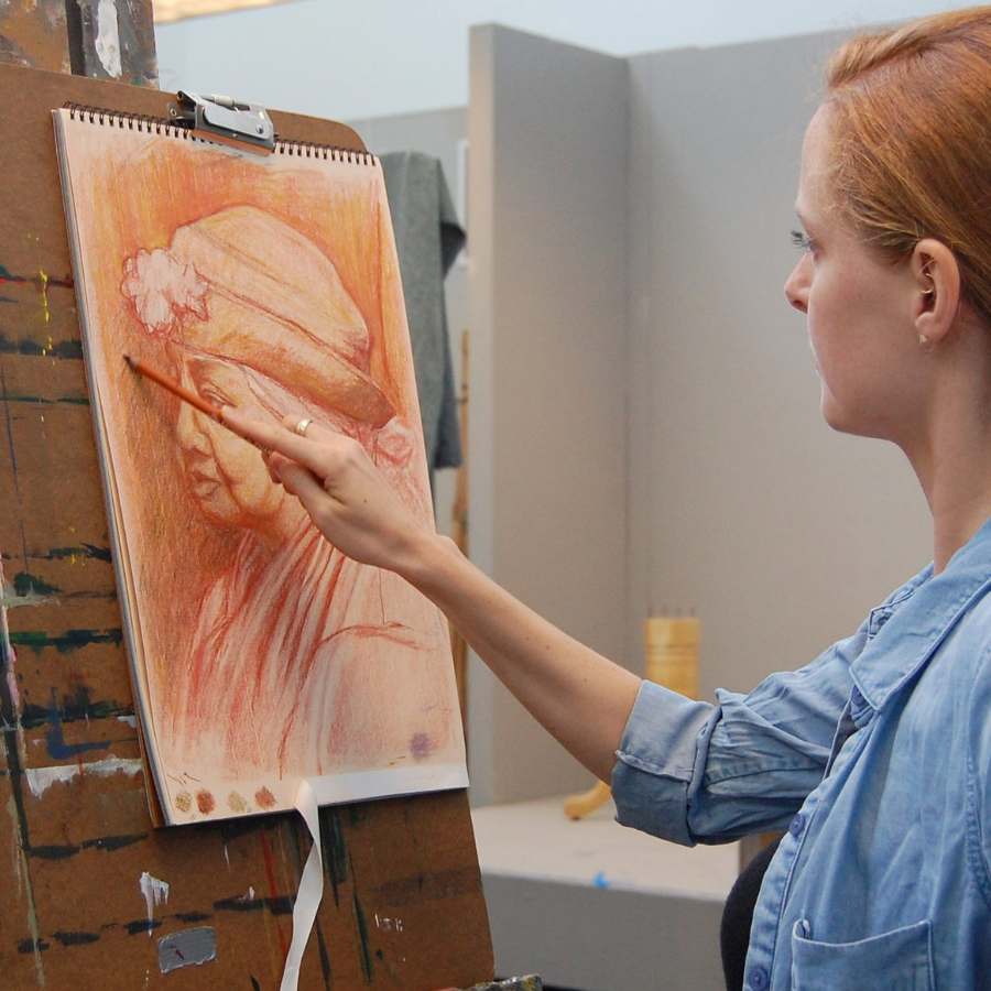 A woman with auburn hair uses a red pastel to sketch a detailed portrait on a spiral-bound pad resting on a wooden easel.