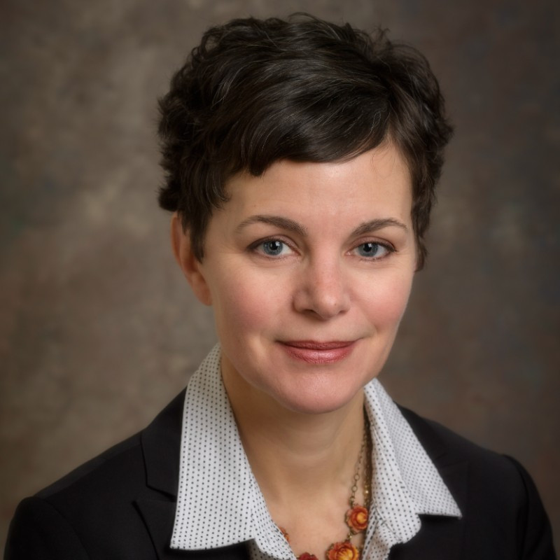 Portrait of a person with short dark hair and a warm smile, wearing a black blazer and a white polka dot shirt, set against a blurred brown background.