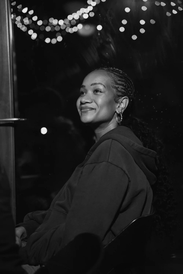 Black and white portrait of a smiling person with braided hair and hoop earrings, sitting in a dimly lit space with blurred string lights behind them.